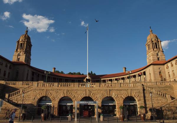 The curved sandstone front of the Union Buildings in Pretoria, with a row of arched entrances beneath a stone terrace, two domed clock towers, a tall flagpole and a bird in flight under a deep blue sky.