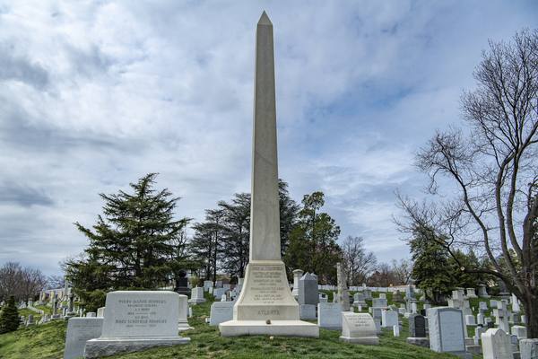 A tall stone obelisk on a grassy hill among rows of gravestones and trees under a cloudy sky.