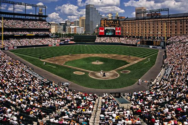 A wide view of Oriole Park at Camden Yards on a sunny day, with a packed grandstand, a striped green field, the brick warehouse beyond right field, the Baltimore skyline and a scoreboard showing the name Cal Ripken.