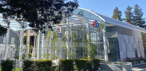 A white-framed glass building with a curved roof and a colorful eBay sign, seen through young trees and a dark pine branch under a blue sky.