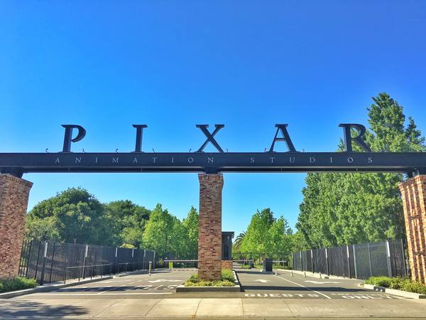 A brick-pillared entrance gate with a dark steel beam carrying the large letters PIXAR above the words Animation Studios, under a deep blue sky with green trees behind and an empty driveway below.