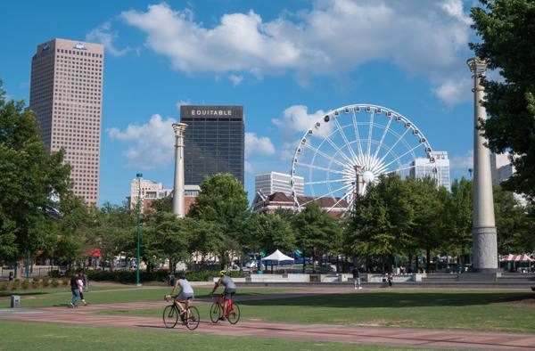 A sunny view across a lawn and a brick path in Centennial Olympic Park, with two cyclists riding along the path, tall office towers and a white Ferris wheel in the distance, and two slender stone towers topped with gold-coloured rings, under a blue sky with white clouds.