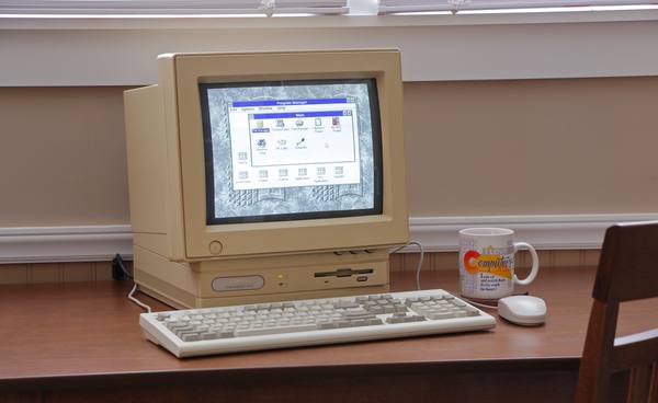 A beige desktop computer with a CRT monitor, a cream keyboard and a mouse on a wooden desk, with an old Windows Program Manager window on the screen.
