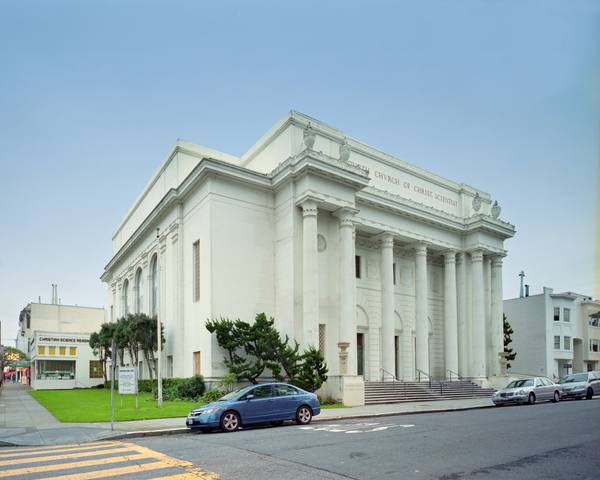 A large white neoclassical building with tall fluted columns across its front, seen from a street corner, with a blue car and other cars parked along the curb.