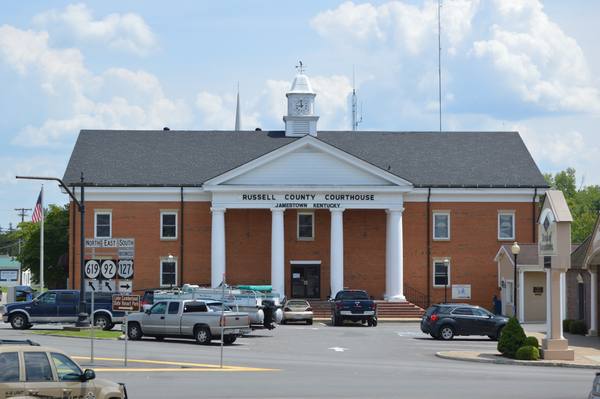 A red-brick county courthouse with a white columned portico and a small clock cupola, with parked pickup trucks and a boat trailer in front under a partly cloudy sky.