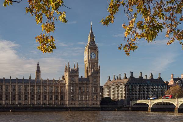 The Palace of Westminster and the Big Ben clock tower seen across the River Thames, with Westminster Bridge at right, framed by autumn leaves under a blue sky.
