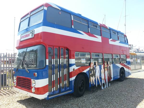A double-decker bus painted as a Union Jack in red, white and blue, with a Spicebus No 19 plate above the windscreen and five life-size cut-out figures of women standing against its side, parked on gravel beside a fence.