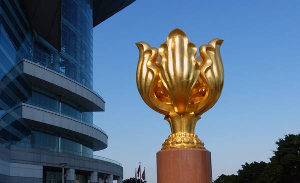 A gold-colored bauhinia flower sculpture on a red granite pedestal beside the curved blue glass walls of a convention centre, against a deep blue sky.