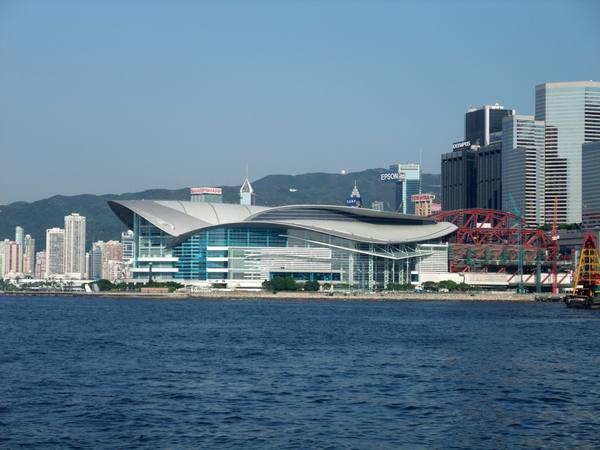 A wave-shaped glass and steel convention centre on the Hong Kong harbour front, seen across blue water with green hills and office towers behind it.