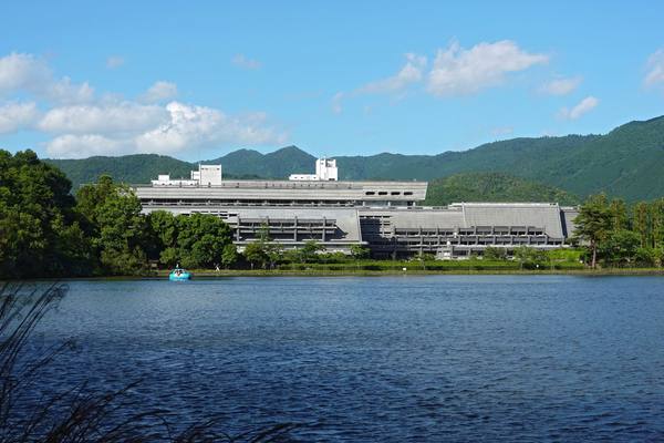 A long, low, grey concrete conference centre set among trees and green hills, seen across a pond under a bright blue sky, with a small blue pedal boat on the water.