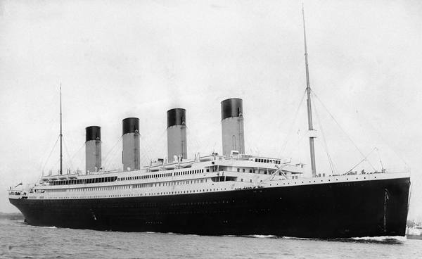 Black-and-white photograph of the four-funnelled ocean liner Titanic under way on calm water, seen from the bow side, with a few tiny figures at the rails.