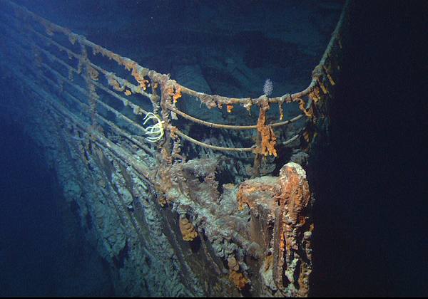 Underwater view of the rust-covered bow railings of the Titanic wreck on the dark seabed, with orange rusticles hanging from the rails and small pale feathery sea creatures beside the railing.