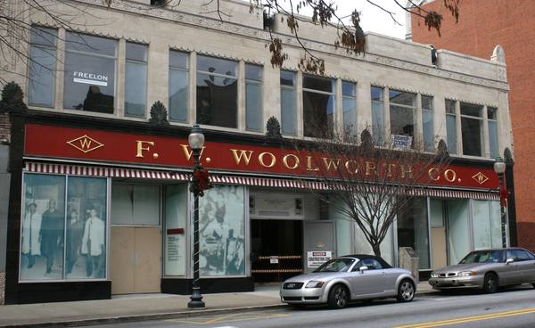 A two-story cream-colored store building with a red F. W. WOOLWORTH CO. sign and striped awning above boarded and glazed shopfronts, with two cars parked on the street in front.