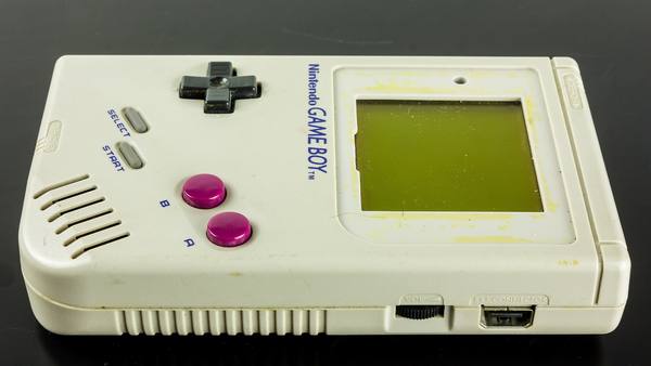 A light gray original Game Boy handheld lying flat on a black surface, showing its cross-shaped D-pad, two magenta buttons, a yellowed screen bezel, and the volume dial and EXT. CONNECTOR port on its bottom edge.