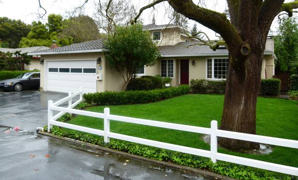 A single-story beige house with a white two-car garage door behind a white fence and green lawn, with a huge moss-covered oak trunk and branches in the right foreground on a wet, overcast day.