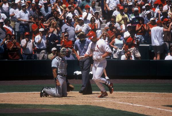 A baseball slugger in a white Cardinals uniform and red helmet trots across home plate past a kneeling catcher and an umpire while fans in the stands behind applaud.