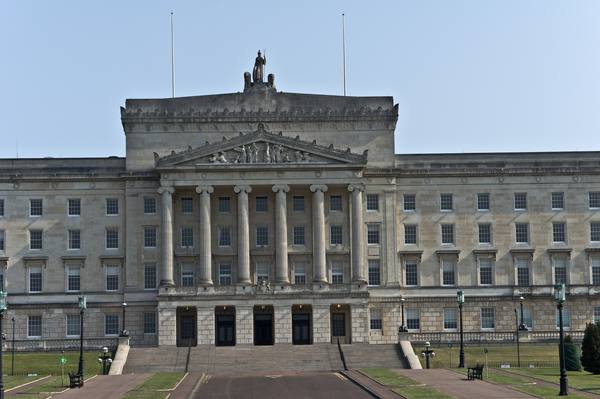 The grey stone neoclassical front of Parliament Buildings at Stormont, with a columned portico, a sculpted pediment, a statue on the roof and a wide ceremonial drive with steps and lawns in front.