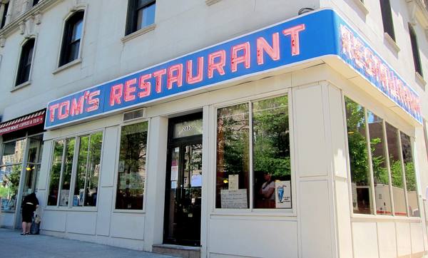 The corner front of a diner with a blue band carrying red neon letters reading Tom's Restaurant, a glass door numbered 2880 and large windows reflecting trees.