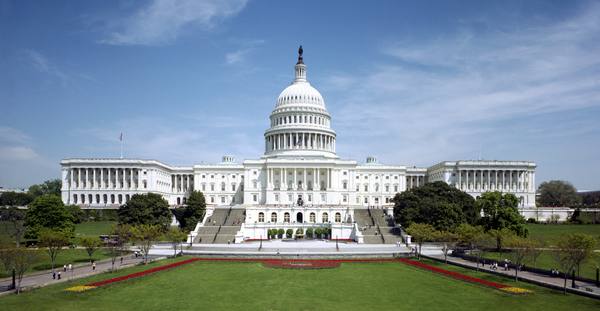 The white-domed U.S. Capitol seen from the west, with its terraced front, a wide green lawn and red and yellow flower beds in the foreground under a blue sky with thin clouds.