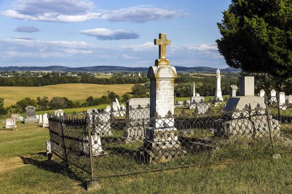 Old headstones and a tall stone cross inside an ornate iron fence on a grassy hillside, with rolling farmland and a blue sky with clouds behind.
