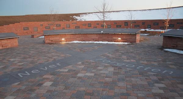 A paved memorial plaza at dusk with a low curved red sandstone wall, a long wall of plaques behind it, patches of snow, bare young trees and a grassy hill, and the words 'Never Forgotten' set into the paving in the foreground.