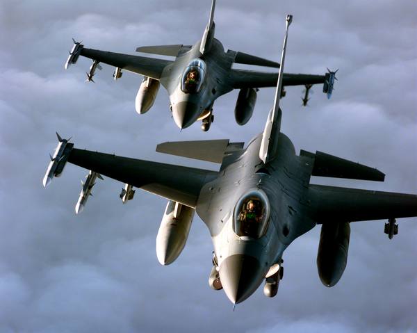 Two grey F-16 fighter jets seen from above in close formation against a cloudy sky, carrying air-to-air missiles and drop tanks.
