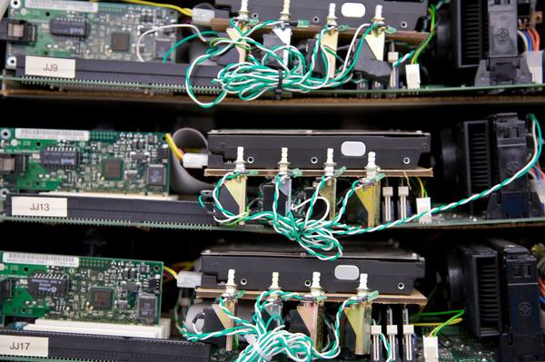 A close view of three bare server trays stacked in a rack, each with a green circuit board and network card, a black hard drive resting on a cork-coloured board and tangled twisted green and white wires, with labels reading JJ9, JJ13 and JJ17.