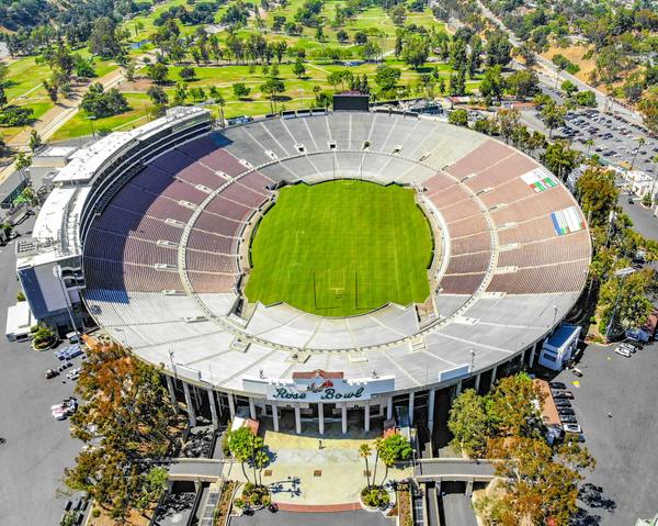 A drone's-eye view of the oval Rose Bowl stadium with its green field, empty tiered seating, the Rose Bowl entrance sign and surrounding trees and parkland.
