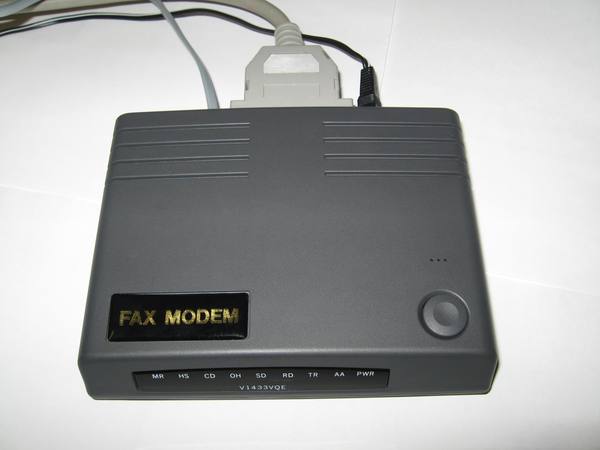 A dark grey external fax modem box with a row of indicator labels along its front edge and a grey serial cable plugged in at the back.