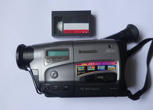 A silver-and-black Panasonic camcorder lying on a pale surface with a small TDK VHS-C videotape cassette above it.