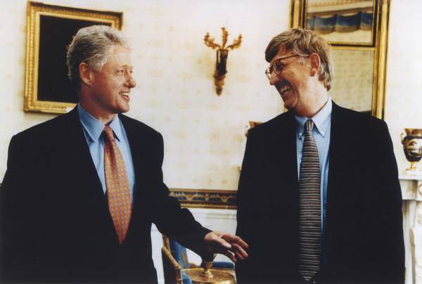 President Bill Clinton, at left, and Francis Collins, at right, smiling and laughing together in a formal room with gilt-framed pictures and a gold wall lamp behind them.