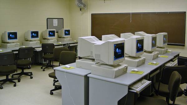 A university computer lab with rows of beige desktop computers and bulky CRT monitors on gray desks, with empty chairs and a blackboard on the wall.