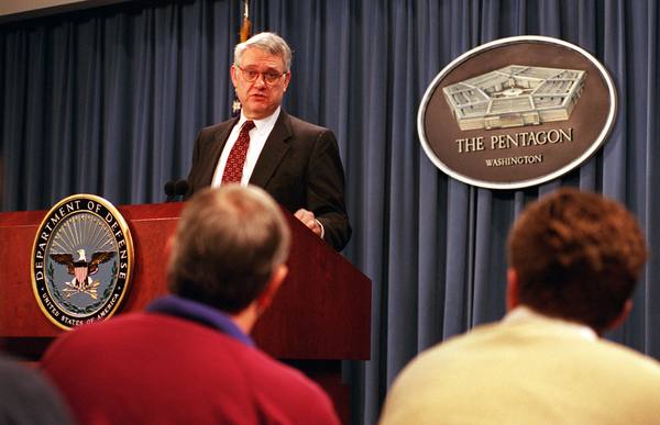 A gray-haired man with glasses, in a dark suit and red tie, speaks at a podium bearing the Department of Defense seal, beside a plaque reading The Pentagon, Washington. Reporters are seen from behind in the foreground.
