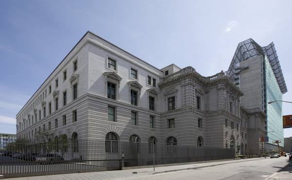 The east facade of a large white granite Beaux Arts courthouse on a street corner behind an iron fence, with a modern glass tower rising behind it under a blue sky.