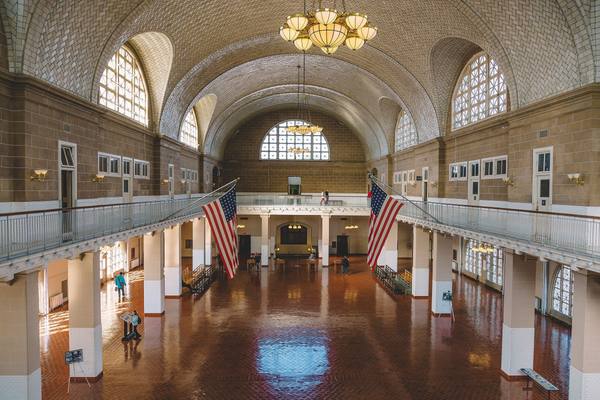 A vast hall with a tiled vaulted ceiling, arched windows, a balcony, two large American flags and a terra-cotta tile floor, with a few visitors far below.