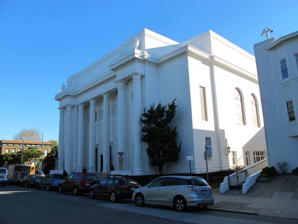 A white building with a row of tall fluted columns across its front stands under a clear blue sky, with parked cars along the street in front of it and a dark evergreen tree beside its side wall.