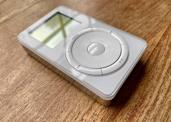 A white first-generation iPod lying at an angle on a wooden table, showing its small screen, a round scroll wheel and a ring of four buttons around it, with the word menu printed at upper left.