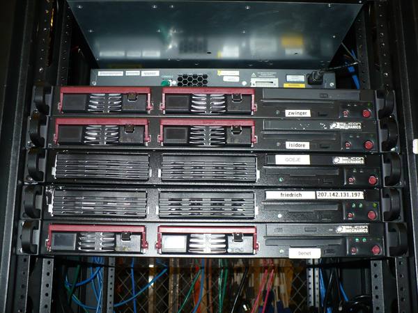Five black rack-mounted servers stacked in a rack, each with a small name label, with a network switch above them and bundles of blue, green and black network cables below.