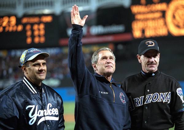 President Bush with one arm raised, standing between a man in a navy Yankees jacket and cap on the left and a man in a black Arizona jersey and cap on the right, with a blurred stadium scoreboard behind them.