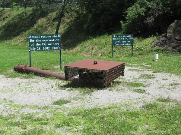 A rusty steel cover with a mesh skirt over a shaft opening on a gravel patch at the foot of a grassy hillside, with a rusty pipe lying on the ground to the left, two green signs behind it and a short white pipe to the right.