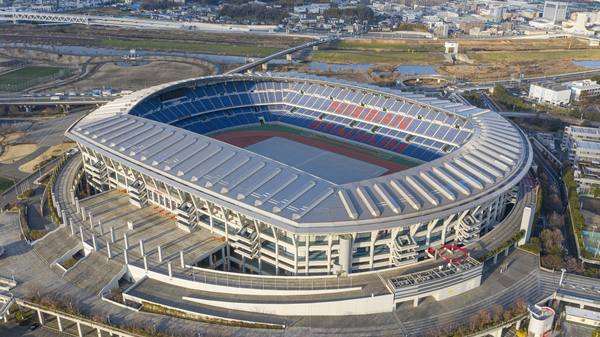 An aerial view of a large oval stadium with a silver roof, blue seats with large red lettering reading Nissan Stadium, a running track and pitch, and surrounding roads and a river.