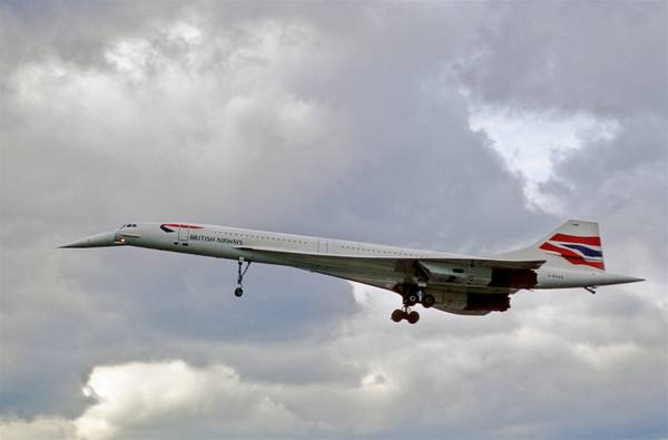 A white Concorde with its nose drooped and landing gear down, in flight against a cloudy gray sky.