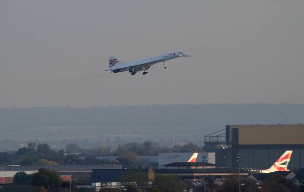 A white British Airways Concorde with its landing gear down, flying low above hangars, trees and British Airways tail fins at Heathrow Airport under a hazy grey sky.