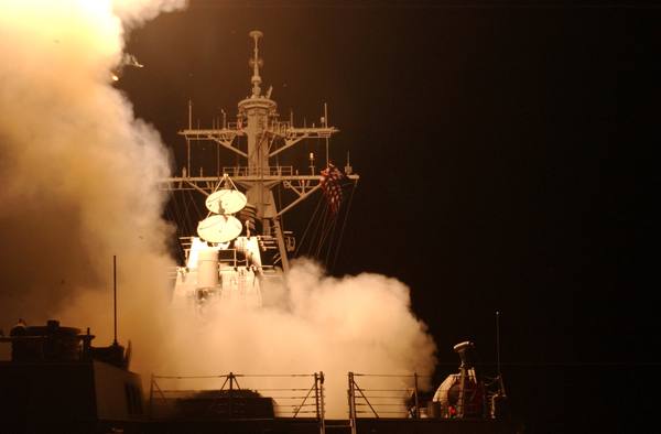 A night view of a destroyer's mast and radar dishes rising from a cloud of glowing smoke after a missile launch, with a U.S. flag flying and a dark sky behind.