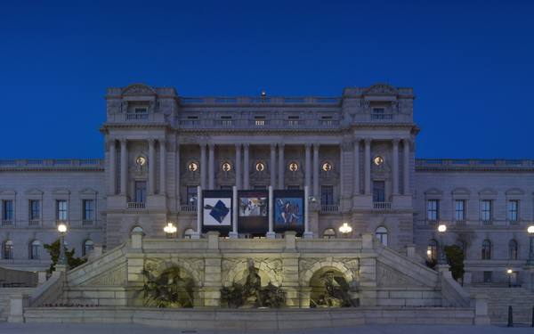 A neoclassical stone library building lit at night under a deep blue sky, with a bronze fountain and staircases in front and banners hanging above the entrance.