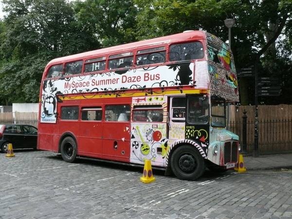 A red double-decker bus with brightly painted patterns on its front and upper deck and a white banner reading "MySpace Summer Daze Bus," parked on a cobbled street beside yellow traffic cones, with trees behind.
