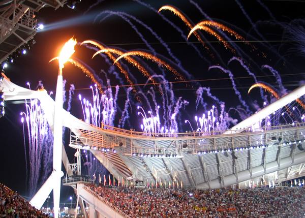 A tall white pole topped by the Olympic flame at left, with orange and violet fireworks arching over the curved white roof structure of the stadium and a packed grandstand below at night.