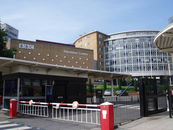 A brick building lettered BBC Television Centre behind a gatehouse and a red-and-white barrier, with a curved glass tower rising behind it under a blue sky.