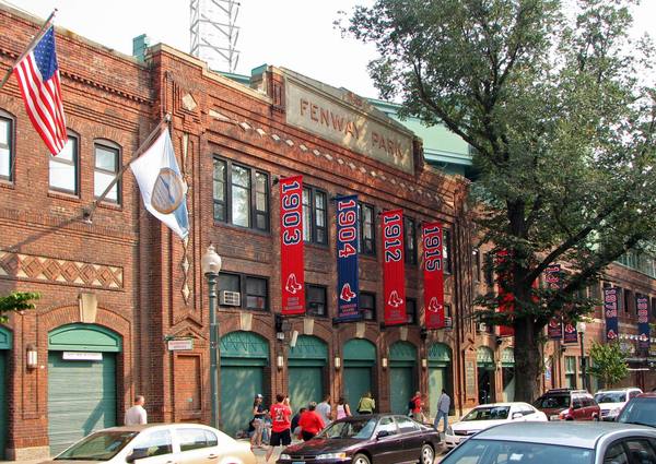 The red brick facade of Fenway Park with its lettered stone sign, an American flag, and red and blue championship banners for 1903, 1904, 1912 and 1915, with a few small figures and parked cars on the street below.