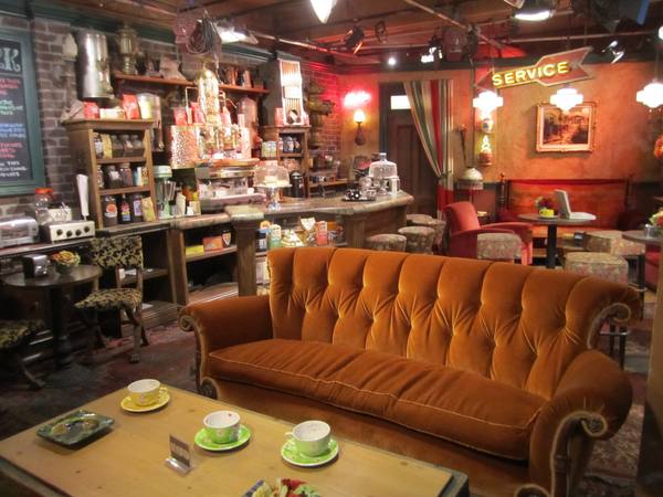 A warmly lit coffeehouse set with a large orange velvet sofa and a wooden coffee table with cups in the foreground, and a counter, espresso machine, shelves of coffee packages and a neon "Service" sign behind.